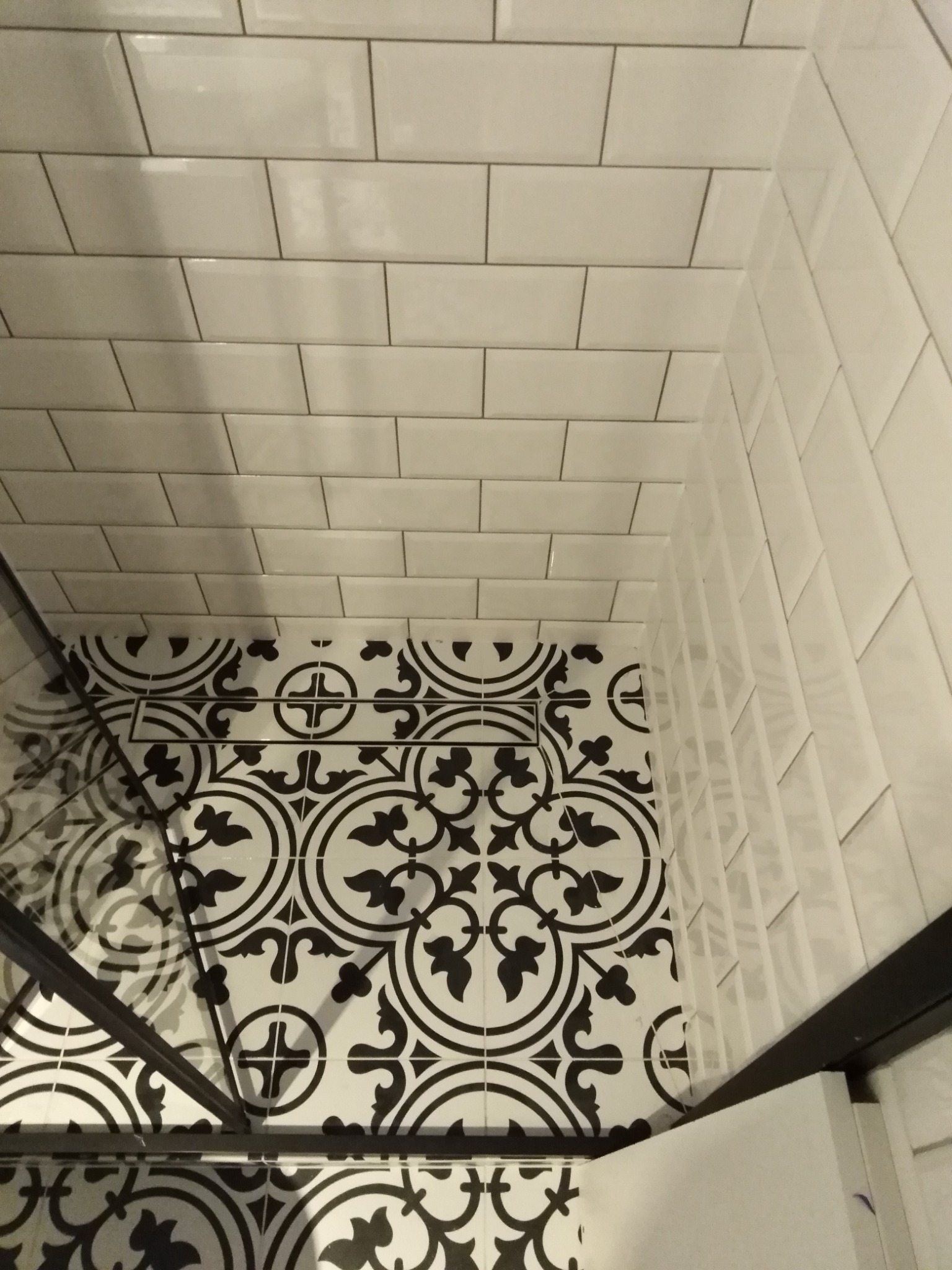 Wykończona kabina prysznicowa z wzorzystą, czarno-białą podłogą i białymi, prostokątnymi płytkami na ścianach ułożonymi w cegiełkę, widoczny odpływ liniowy.