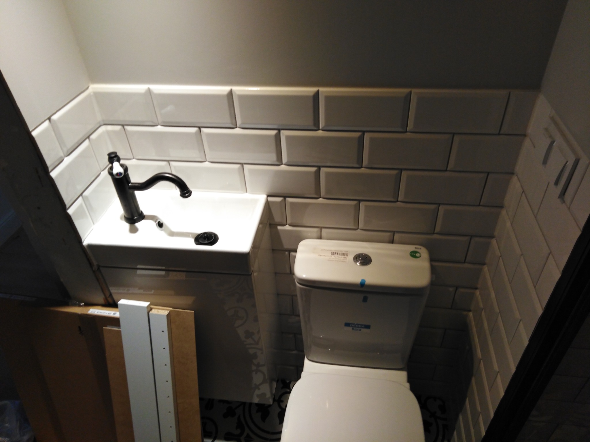 Mała łazienka w trakcie wykańczania: białe, prostokątne płytki na ścianach, umywalka z czarną baterią i toaleta. Na podłodze widoczny fragment wzorzystych płytek. Z lewej strony leżą elementy...