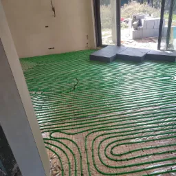 Rozłożone zielone rury ogrzewania podłogowego na srebrnej folii, widoczny fragment wylewki, okno przesuwne i materiały budowlane na zewnątrz.