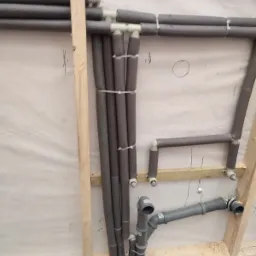 Surowe ujęcie fragmentu instalacji hydraulicznej w trakcie montażu: rury w izolacji termicznej przymocowane do drewnianej konstrukcji ściany, widoczne przyłącza kanalizacyjne i drobne ścinki drewna...