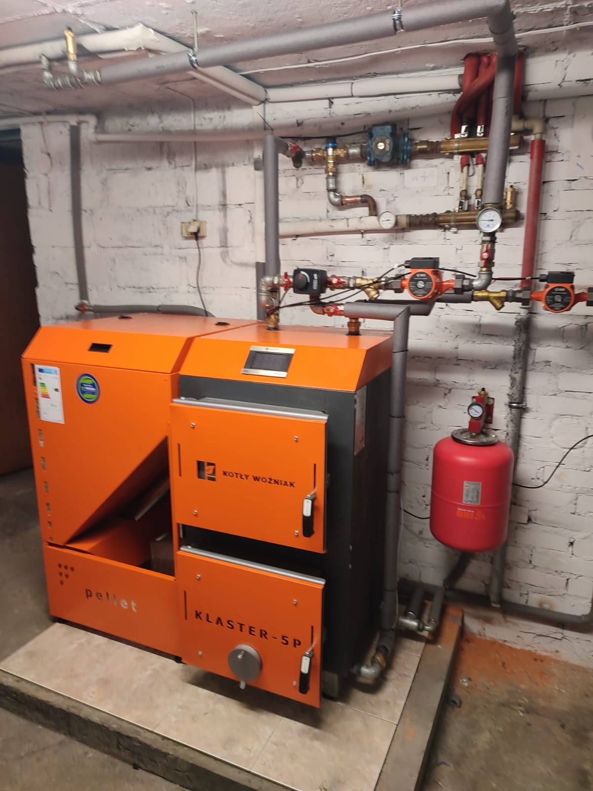 Pomarańczowy piec na pellet marki Kotły Woźniak, model Klaster-5P, zainstalowany w kotłowni z widocznymi rurami, pompami obiegowymi i czerwonym zbiornikiem wyrównawczym.
