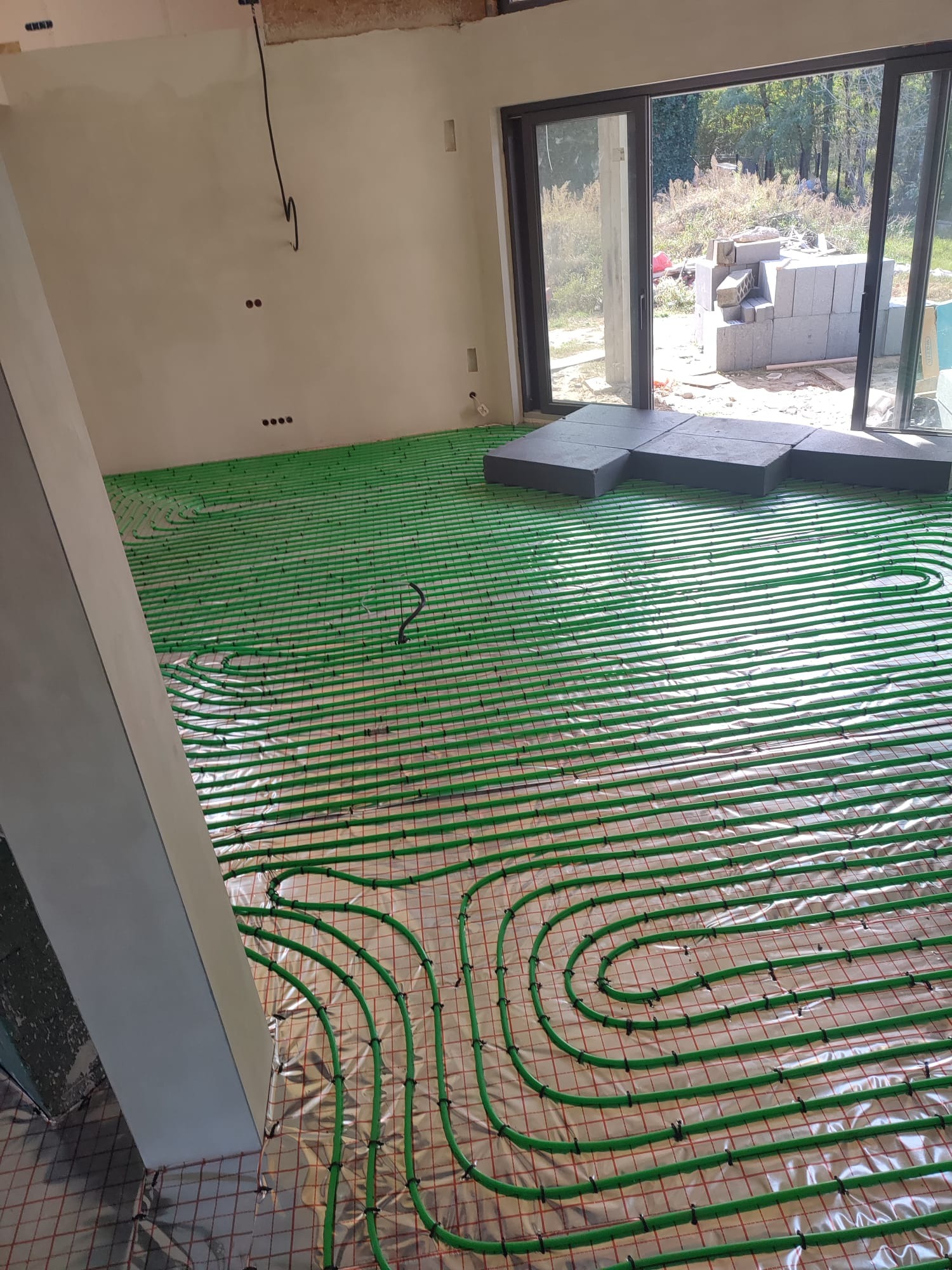 Rozłożone zielone rury ogrzewania podłogowego na srebrnej folii, widoczny fragment wylewki, okno przesuwne i materiały budowlane na zewnątrz.