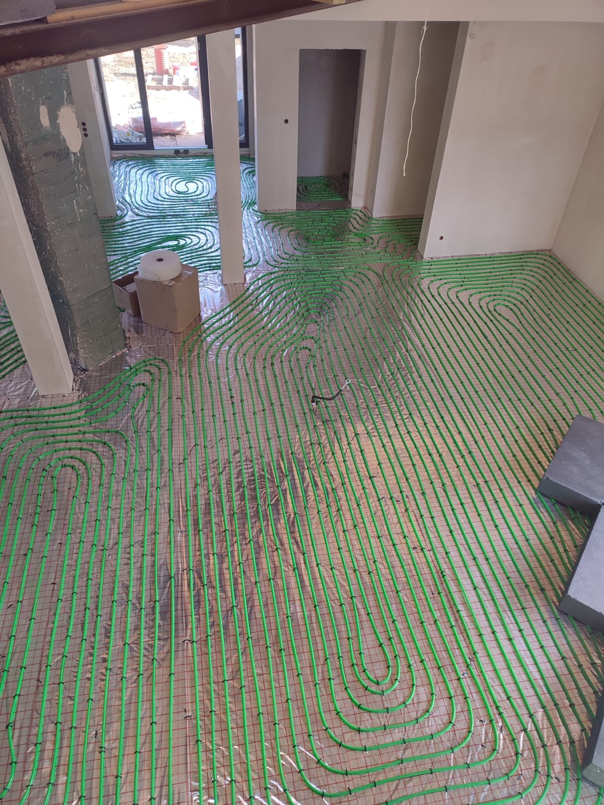 Widok z góry na rozłożone, zielone rurki ogrzewania podłogowego na folii izolacyjnej z czerwoną siatką w trakcie montażu w surowym wnętrzu budynku.