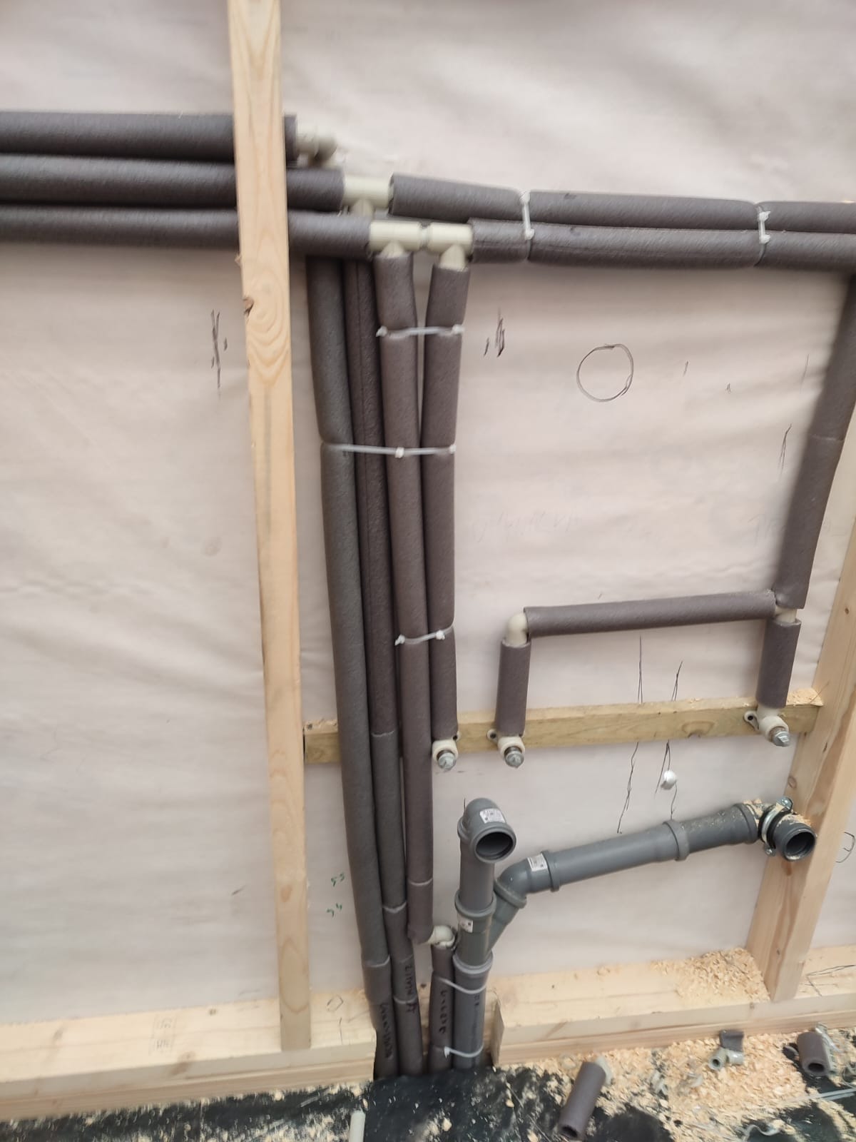 Surowe ujęcie fragmentu instalacji hydraulicznej w trakcie montażu: rury w izolacji termicznej przymocowane do drewnianej konstrukcji ściany, widoczne przyłącza kanalizacyjne i drobne ścinki drewna...