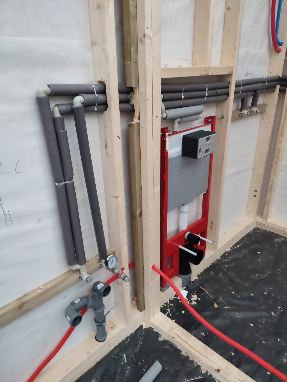 Wnętrze w trakcie prac instalacyjnych: widoczne rury grzewcze zaizolowane szarą pianką, czerwone rury instalacji wodnej oraz stelaż podtynkowy WC z czarnym modułem, wszystko zamontowane...