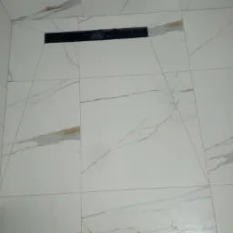 Białe płytki podłogowe imitujące marmur z czarnym odpływem liniowym w kabinie prysznicowej, widoczny fragment buta.