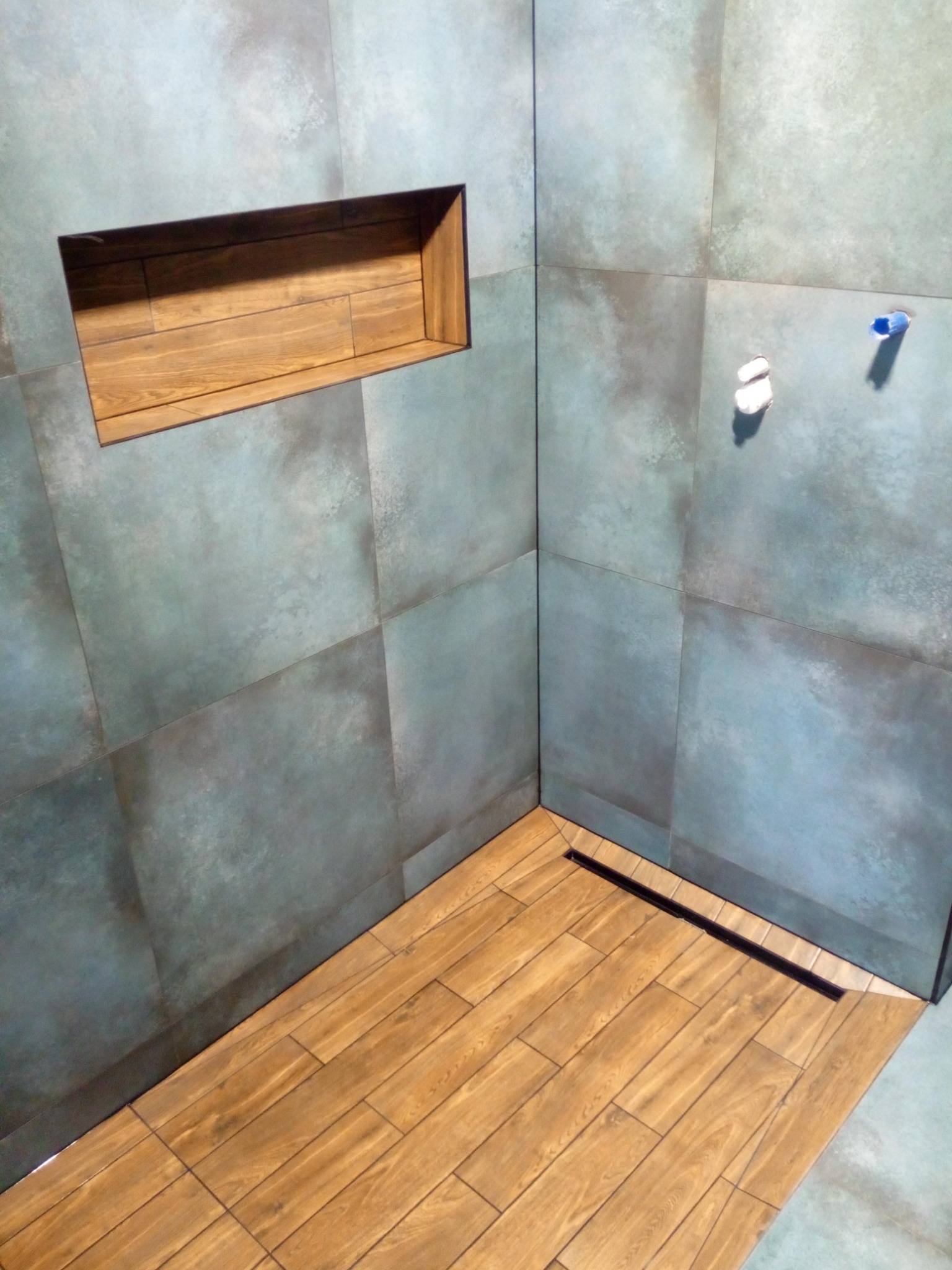 Nowoczesna kabina prysznicowa z szaro-niebieskimi płytkami imitującymi beton i drewnianą podłogą oraz wnęką w ścianie wykończoną drewnem.
