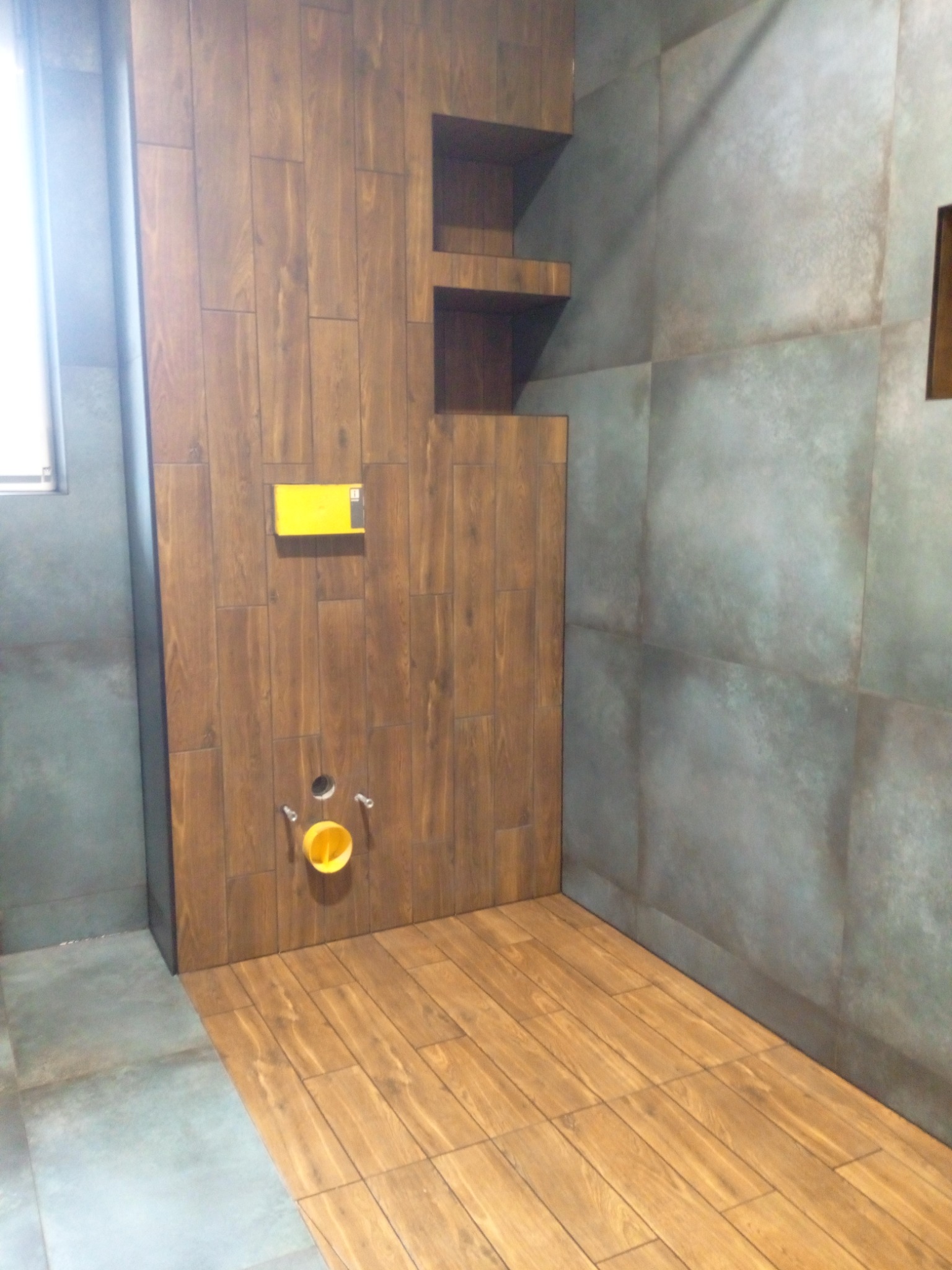Wykończona łazienka z drewnopodobnymi płytkami na ścianie i podłodze, widoczny stelaż podtynkowy WC z żółtą osłoną i fragment półek w zabudowie.