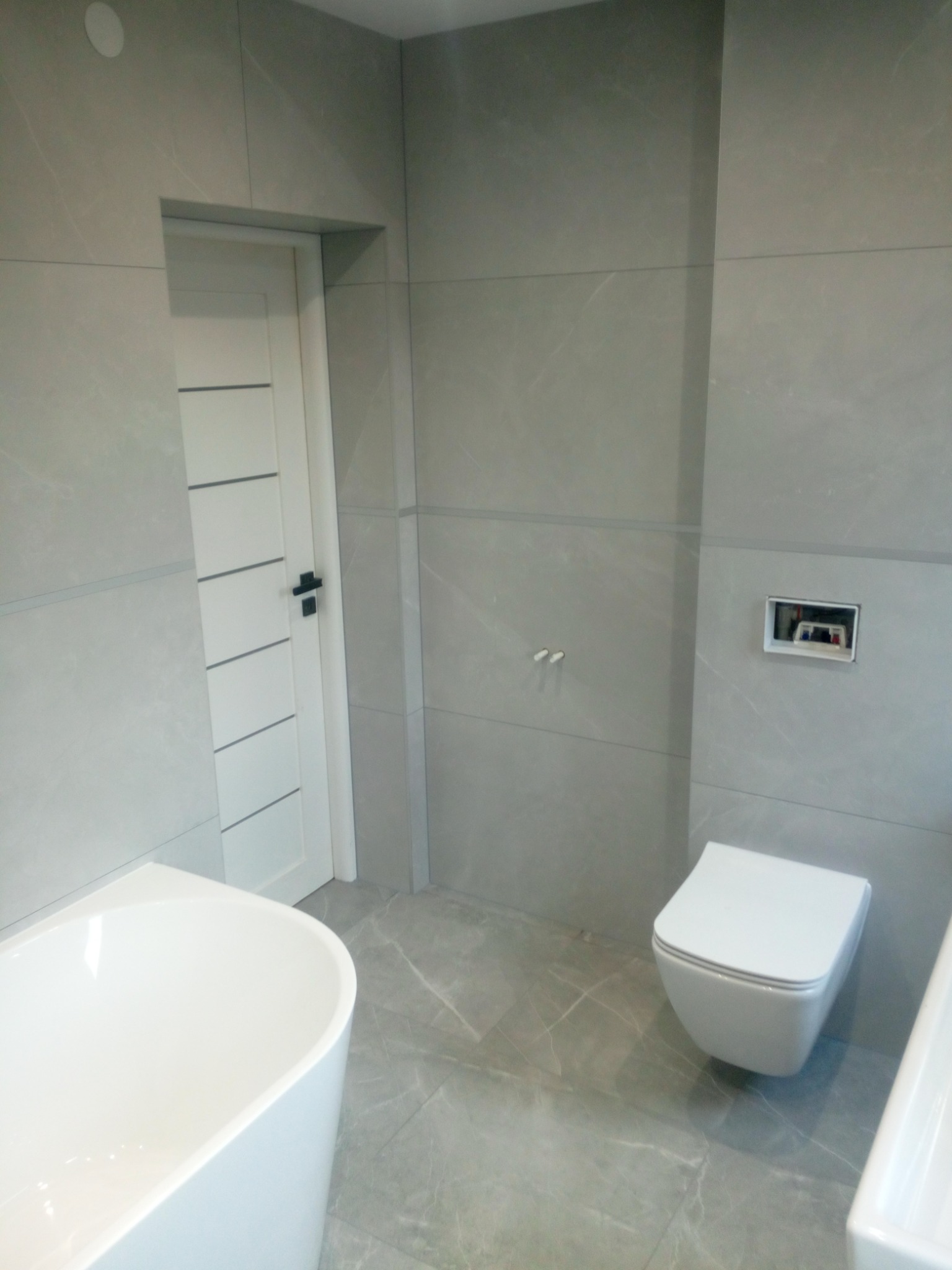 Nowoczesna łazienka z szarymi płytkami imitującymi marmur, białą wanną wolnostojącą, podwieszaną toaletą i białą, minimalistyczną stolarką drzwiową z czarną klamką.