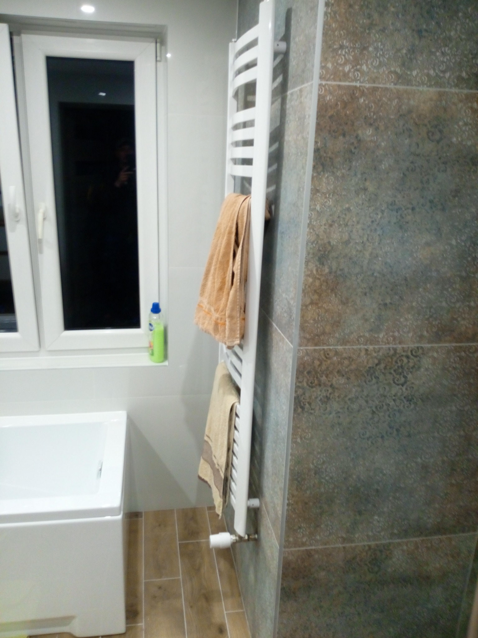 Nowoczesna łazienka z wanną, grzejnikiem drabinkowym z ręcznikami i dekoracyjnymi płytkami ściennymi w odcieniach brązu i szarości, widok z boku