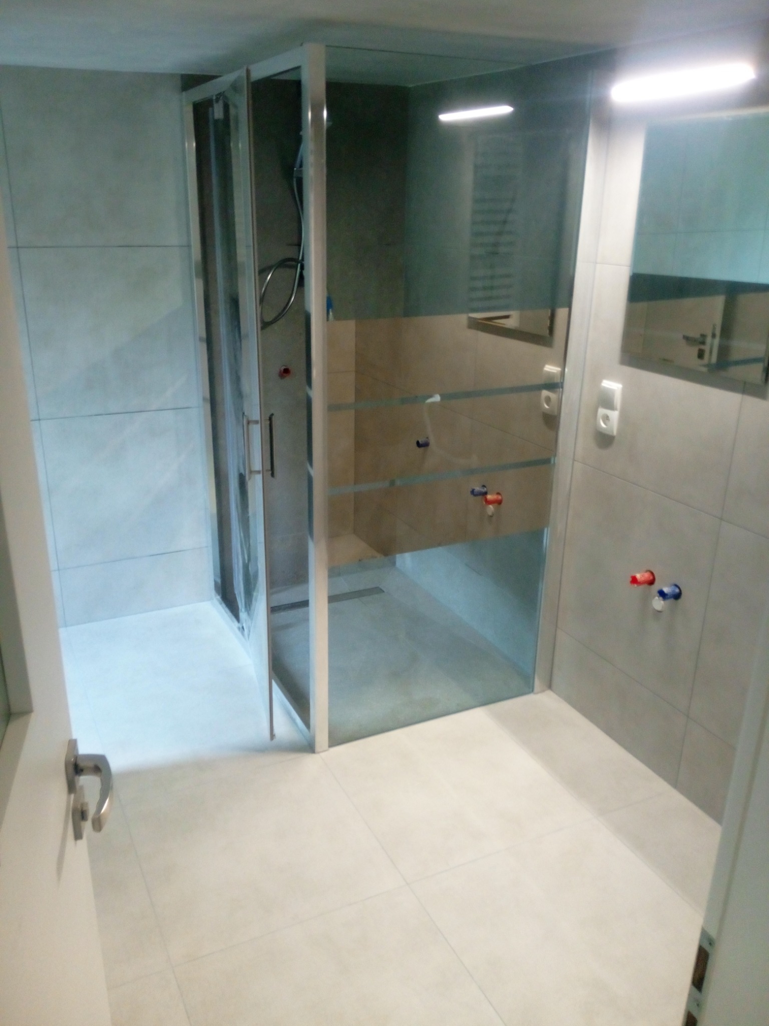 Nowoczesna kabina prysznicowa z przezroczystego szkła, zamontowana w wykafelkowanej łazience z jasnymi płytkami, widoczne baterie i lustro.