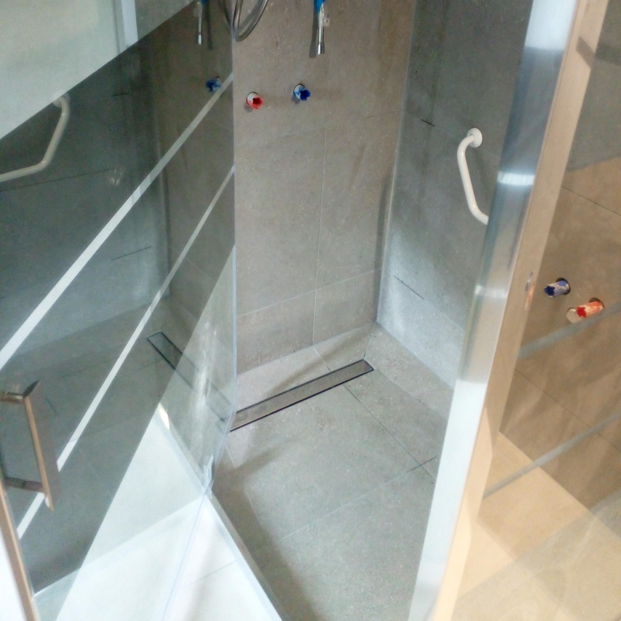 Nowoczesna kabina prysznicowa z odpływem liniowym, szare płytki na ścianach i podłodze, z widocznymi przyłączami wody i uchwytem dla niepełnosprawnych.