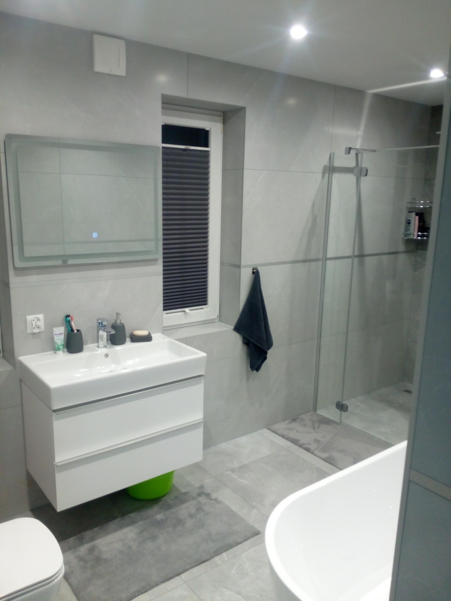 Nowoczesna łazienka w odcieniach szarości z białą umywalką na szafce, szklaną kabiną prysznicową i roletą w oknie, akcentem kolorystycznym jest zielony pojemnik pod umywalką.