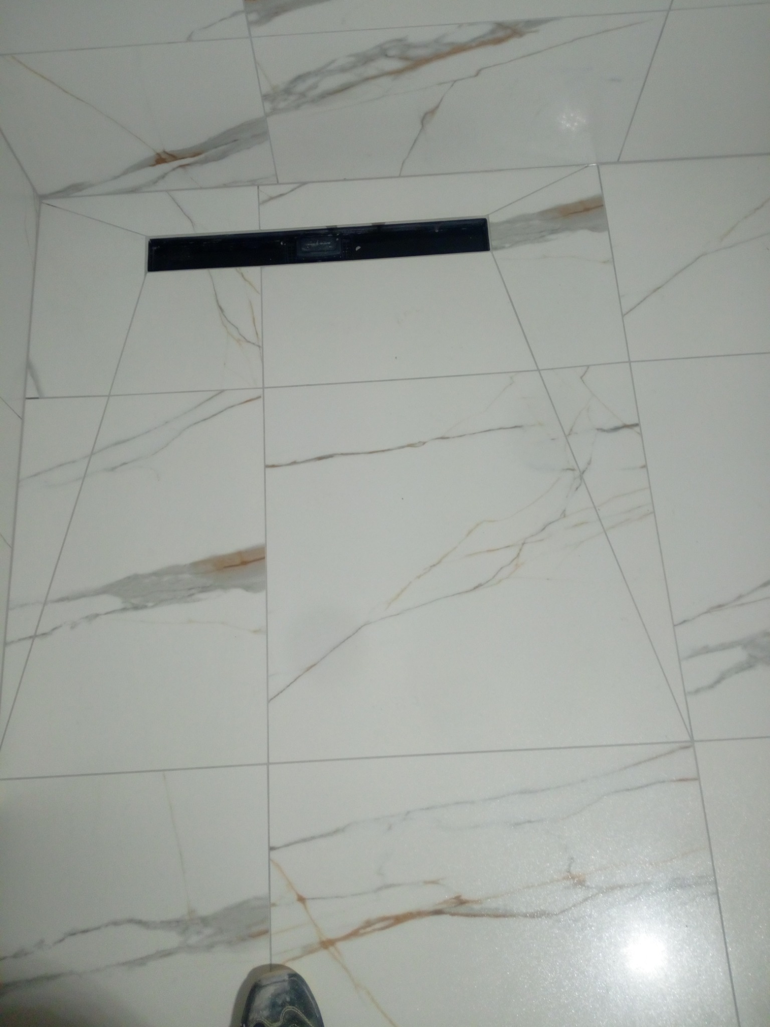 Białe płytki podłogowe imitujące marmur z czarnym odpływem liniowym w kabinie prysznicowej, widoczny fragment buta.