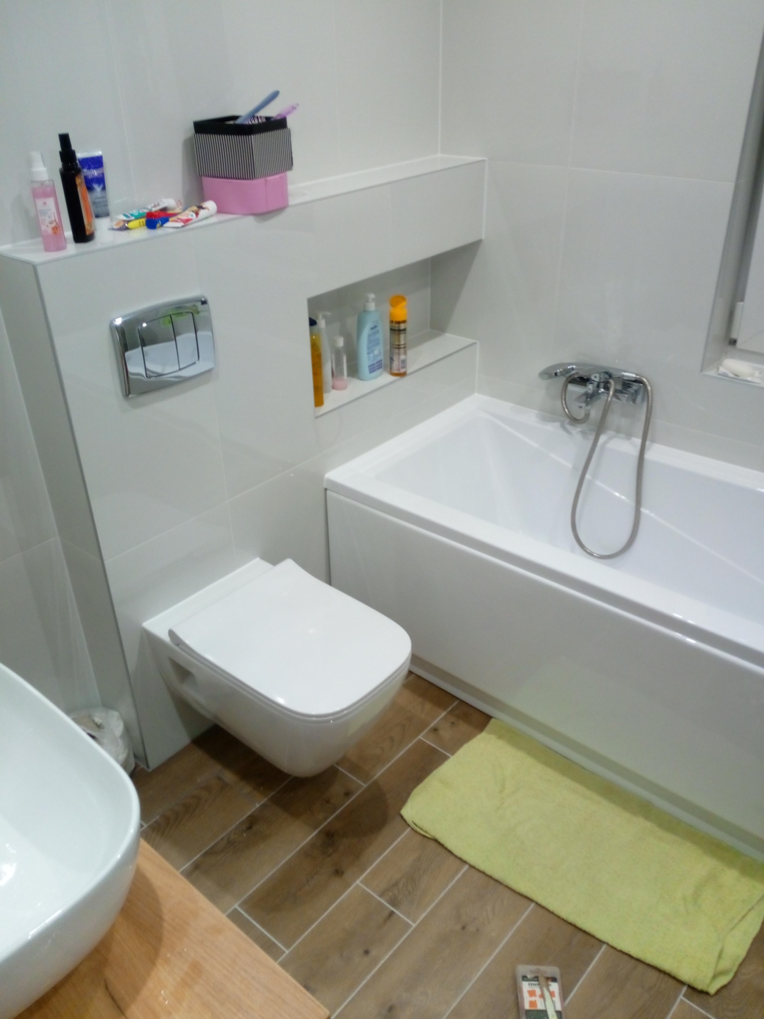 Nowoczesna łazienka z podwieszaną toaletą, białą wanną z chromowaną baterią i prysznicem, drewnopodobną podłogą i zielonym ręcznikiem.