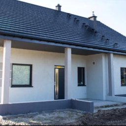Przemo - Nowoczesny dom murowany z grafitowym dachem i jasną elewacją, z elementami dekoracyjnymi w postaci poziomych pasów, widok z zewnątrz na front budynku.
