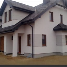 Przemo - Nowoczesny dom jednorodzinny z szarą dachówką i jasną elewacją, widoczne rynny i okna z brązowymi ramami, w tle inne budynki.