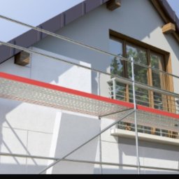 Przemo - Dom jednorodzinny w trakcie prac elewacyjnych, widoczne rusztowanie z czerwonymi elementami, ściana w jasnym kolorze, okno z drewnianą ramą.