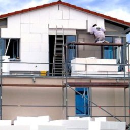 Przemo - Dwupiętrowy dom w trakcie nakładania białych płyt izolacyjnych na elewację, widoczne rusztowania i drabina, pracownik w białym kombinezonie przy pracy.
