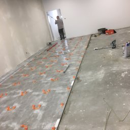 Przemo - Układanie szarych płytek podłogowych z pomarańczowymi krzyżykami dystansowymi; pracownik maluje ścianę w tle, widoczne narzędzia i poziomica.