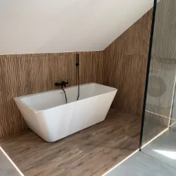 Nowoczesna łazienka na poddaszu z wolnostojącą białą wanną, czarną armaturą, ścianą wykończoną drewnopodobnymi lamelami z ukrytym oświetleniem LED i kabiną prysznicową ze szklaną ścianą.