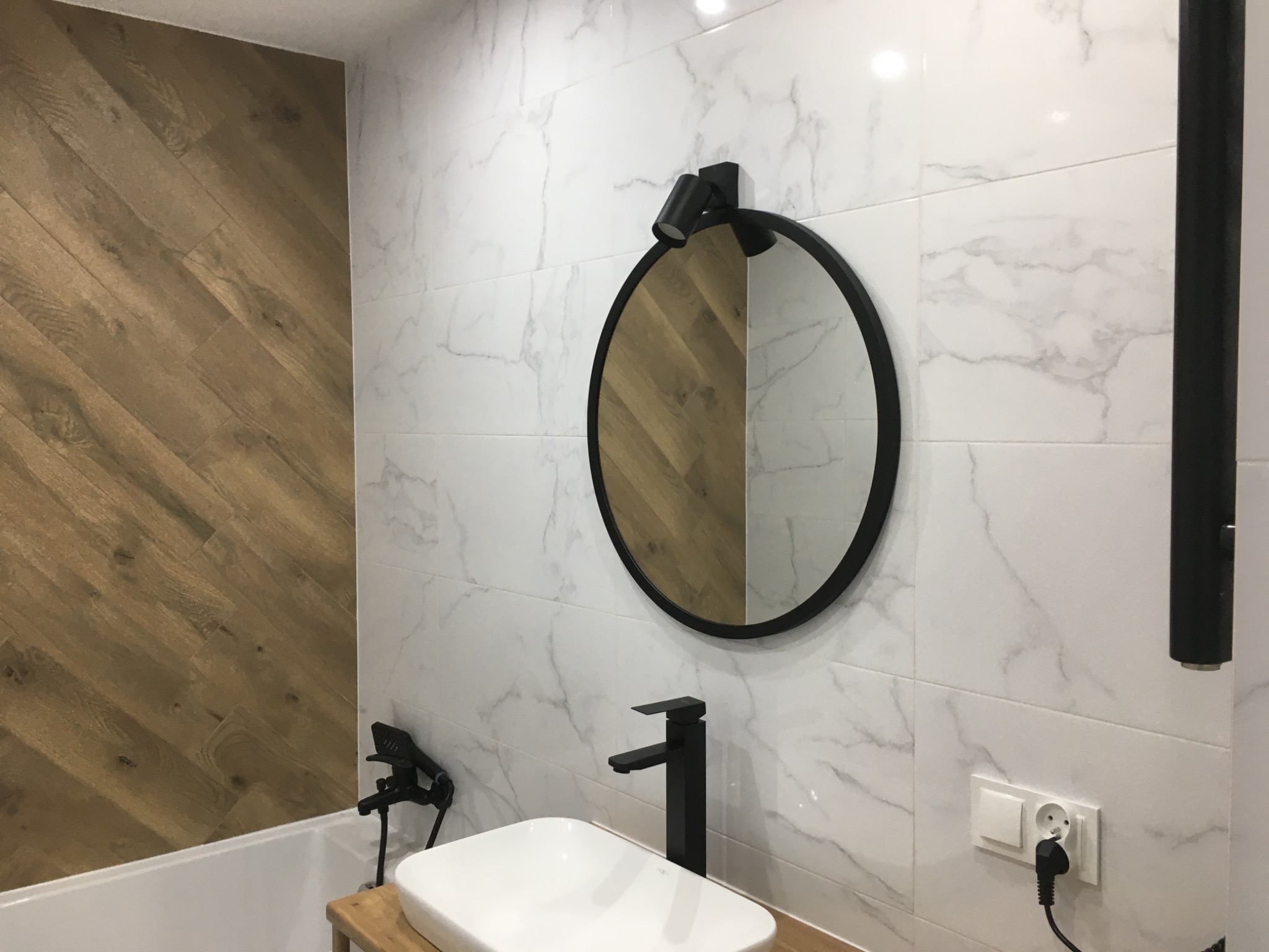 Nowoczesna łazienka z białymi marmurkowymi płytkami, okrągłym lustrem z czarną ramą i punktowym oświetleniem, białą umywalką na drewnianym blacie i czarną armaturą, drewnopodobną ścianą w jodełkę...