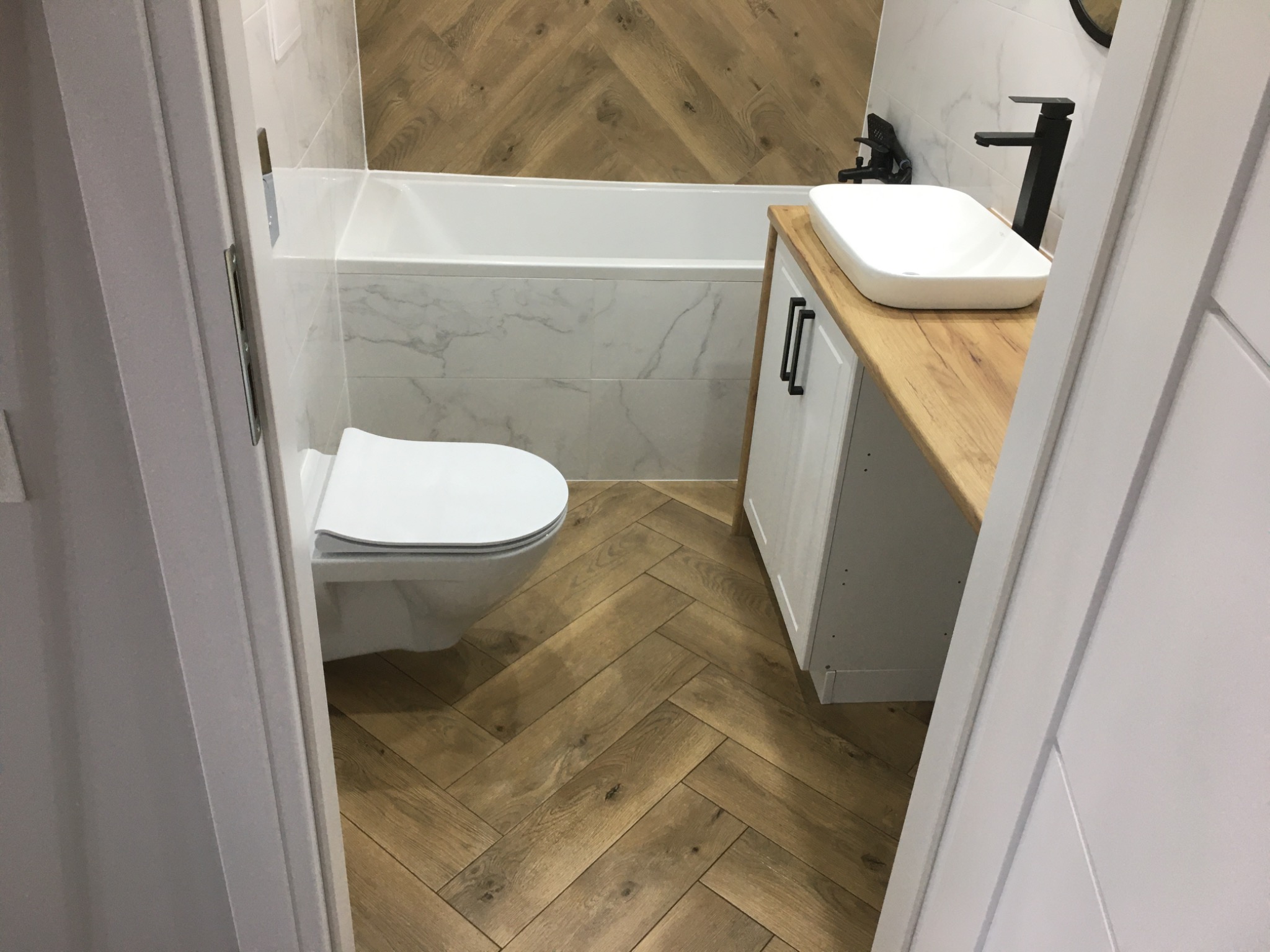 Nowoczesna łazienka z drewnopodobną podłogą ułożoną w jodełkę, białą armaturą i marmurowymi płytkami na ścianach, widziana zza uchylonych drzwi.