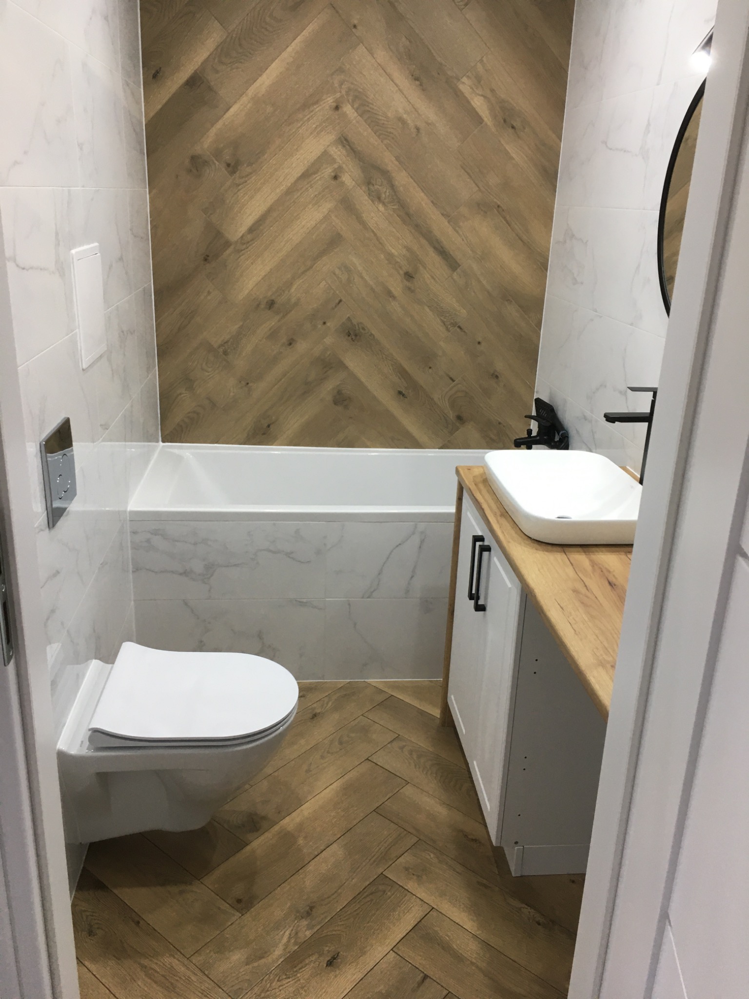 Nowoczesna łazienka z drewnopodobną okładziną w jodełkę na podłodze i ścianie, biała armatura i umywalka nablatowa na drewnianym blacie.