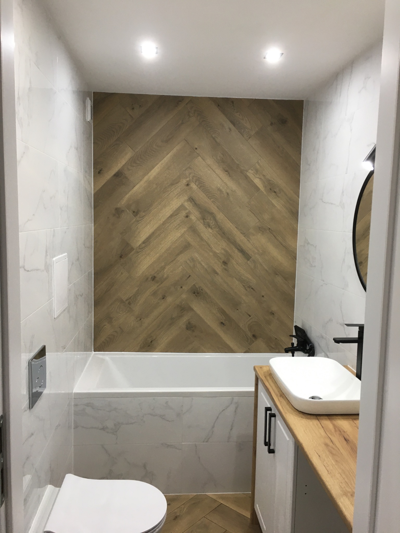 Nowoczesna łazienka z drewnianą ścianą w jodełkę, białą armaturą i marmurowymi płytkami, oświetlona punktowo.