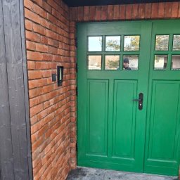 Spec Tynk Paweł Zawrotny - Ceglana ściana z zielonymi drzwiami z szybkami, odbicie osoby w szybie. Nowoczesne wykończenie zewnętrzne budynku z elementami drewna i kamienia.