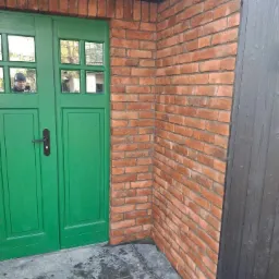 Ceglana ściana przy wejściu z zielonymi drzwiami, odbicie osoby robiącej zdjęcie w szybie. Szara, kamienna posadzka i czarna drewniana kolumna.