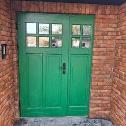 Spec Tynk Paweł Zawrotny - Świeżo pomalowane na zielono dwuskrzydłowe drzwi wejściowe z szybkami, otoczone czerwoną cegłą. W szybach odbija się osoba robiąca zdjęcie.