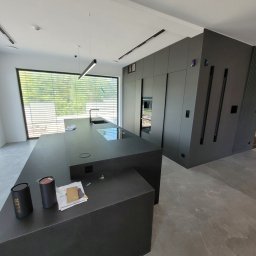 Spec Tynk Paweł Zawrotny - Nowoczesna, minimalistyczna kuchnia w odcieniach szarości z wyspą kuchenną i dużym oknem z żaluzjami, oświetlona lampami liniowymi. Widoczne detale wykończenia.