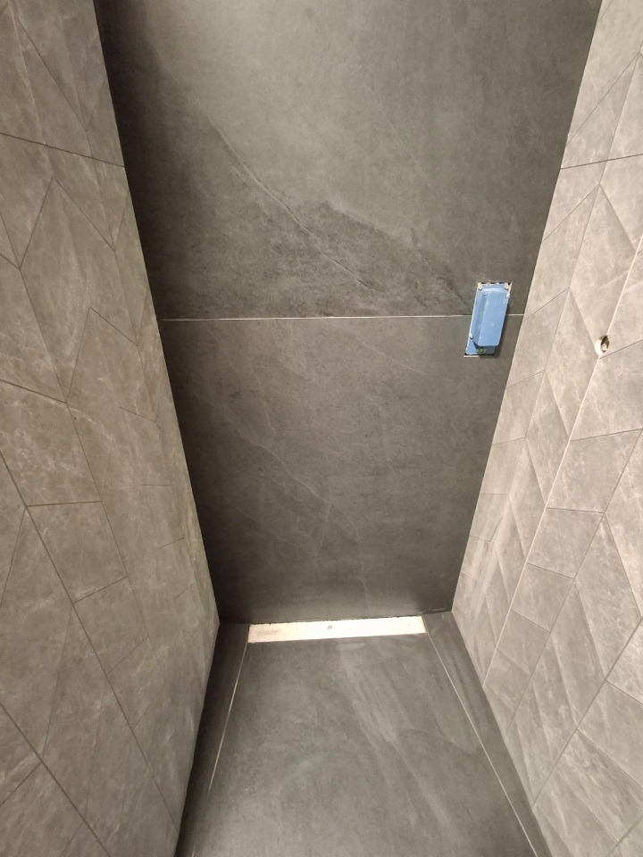 Nowoczesna łazienka z szarymi, dużymi płytkami na ścianach i podłodze. Geometryczny wzór na jednej ze ścian, widoczny odpływ liniowy w podłodze.