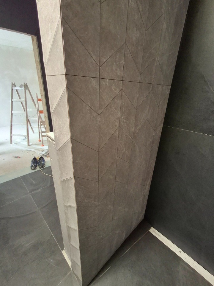 Wykończona ściana z płytek w odcieniach szarości z geometrycznym wzorem, widoczny fragment podłogi z płytek i drabina w tle, wnętrze w trakcie remontu.