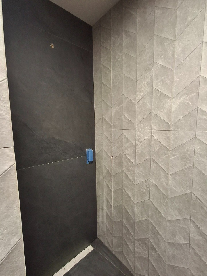 Nowoczesna łazienka po remoncie: ściany wyłożone szarymi płytkami w jodełkę i ciemnymi płytami imitującymi kamień. Widoczna puszka elektryczna.