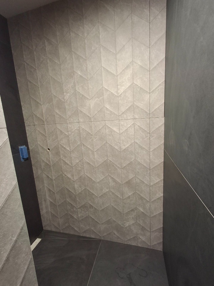 Ściana i podłoga wyłożone szarymi, wzorzystymi płytkami w jodełkę. Widoczny fragment ściany z gładkich, ciemniejszych płytek. Minimalistyczny design łazienki.