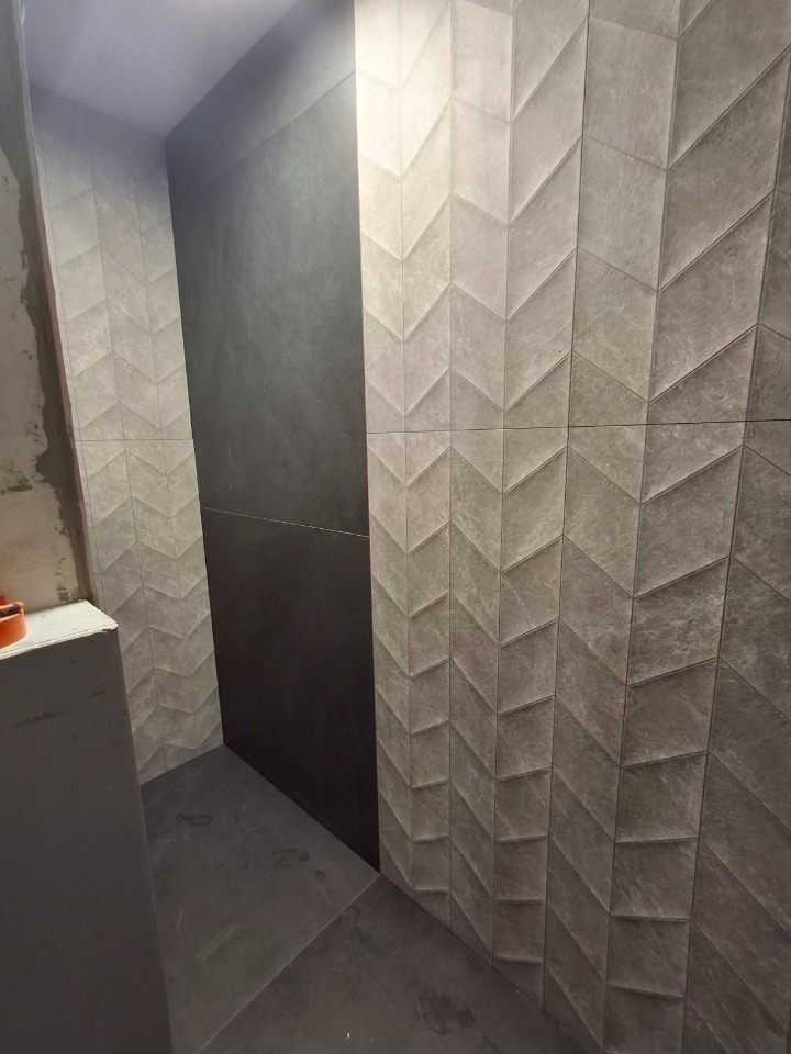 Nowoczesna łazienka z szarymi płytkami ułożonymi w jodełkę na ścianie, kontrastującymi z gładką, ciemną płytą. Minimalistyczny design i staranne wykończenie.