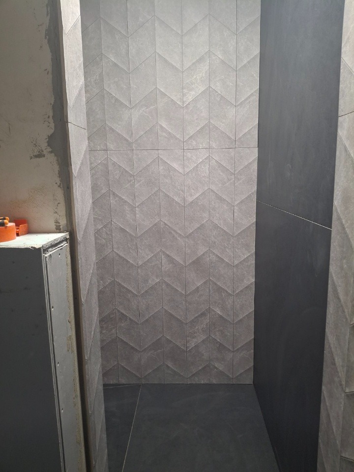 Wykończona ściana w łazience z szarymi płytkami ułożonymi we wzór chevron. Częściowo widoczna podłoga z ciemnych płytek. Minimalistyczny design.