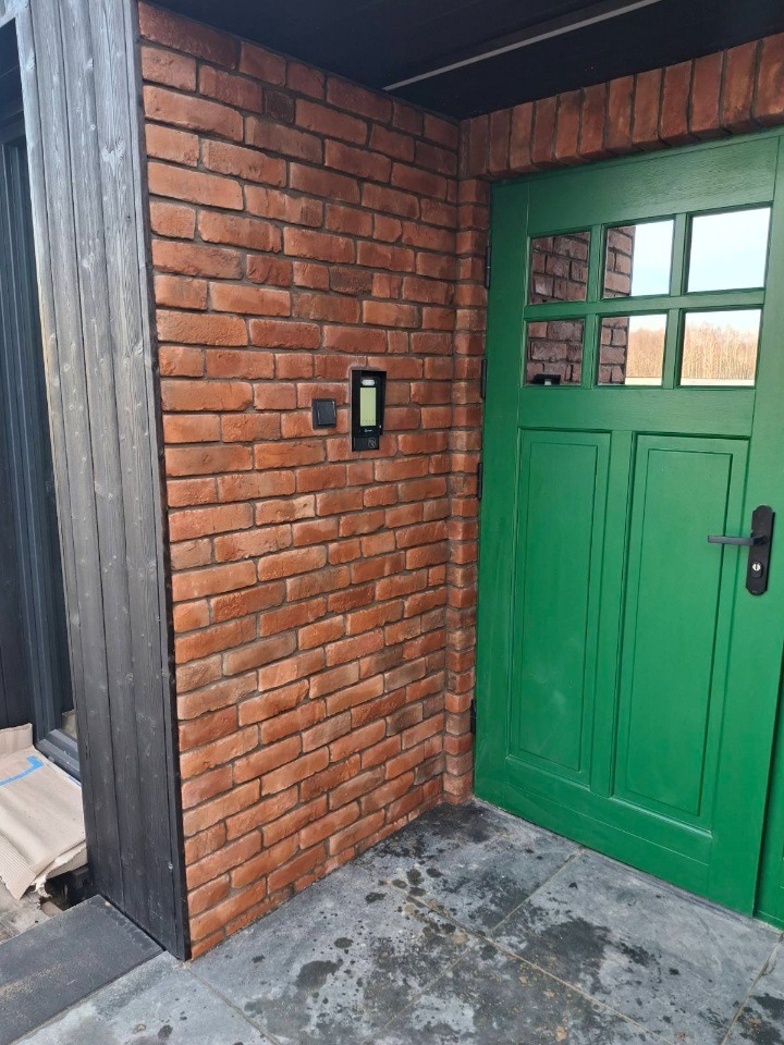 Eleganckie wykończenie wejścia: ceglana ściana z domofonem, zielone drzwi z szybkami i grafitowa podłoga z kamienia. Detale wykończeniowe.