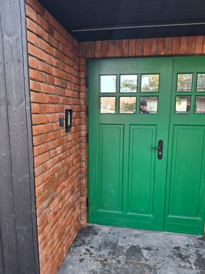 Ceglana ściana z zielonymi drzwiami z szybkami, odbicie osoby w szybie. Nowoczesne wykończenie zewnętrzne budynku z elementami drewna i kamienia.