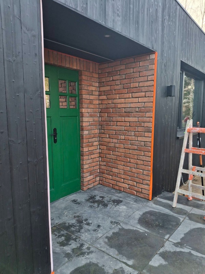 Wykończenie elewacji cegłą klinkierową i drewnem w Warszawie. Zielone drzwi wejściowe, czarne deski elewacyjne. Widoczna drabina.