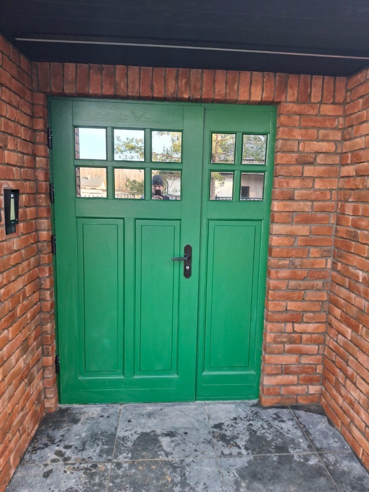 Świeżo pomalowane na zielono dwuskrzydłowe drzwi wejściowe z szybkami, otoczone czerwoną cegłą. W szybach odbija się osoba robiąca zdjęcie.