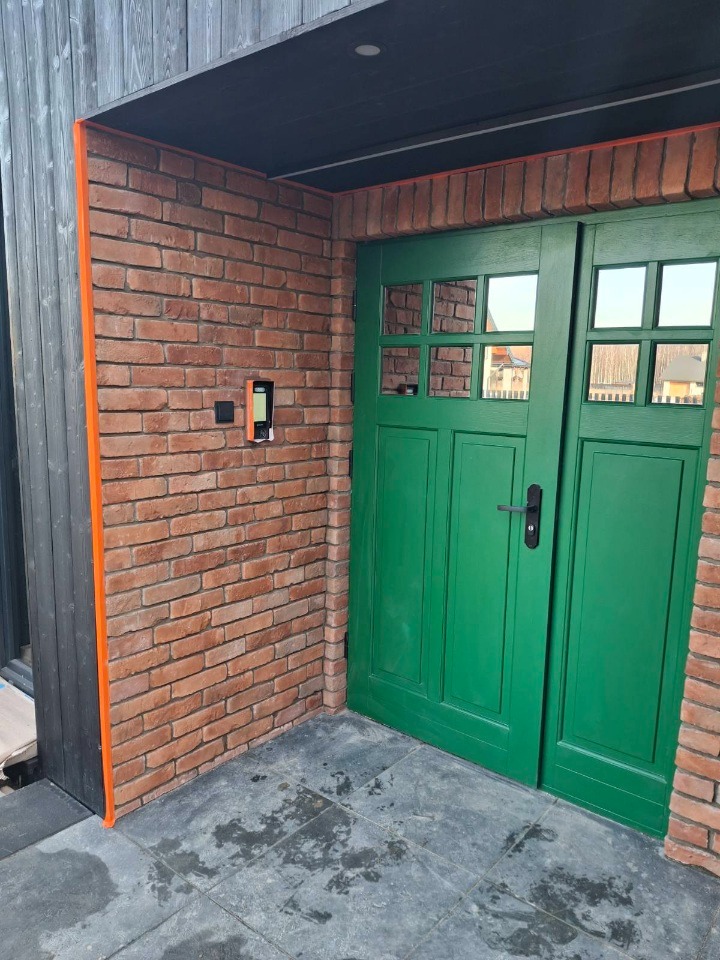 Wejście do domu z ceglaną elewacją i zielonymi drzwiami z szybkami. Widoczny domofon i szara podłoga z płytek. Kątowe ujęcie.
