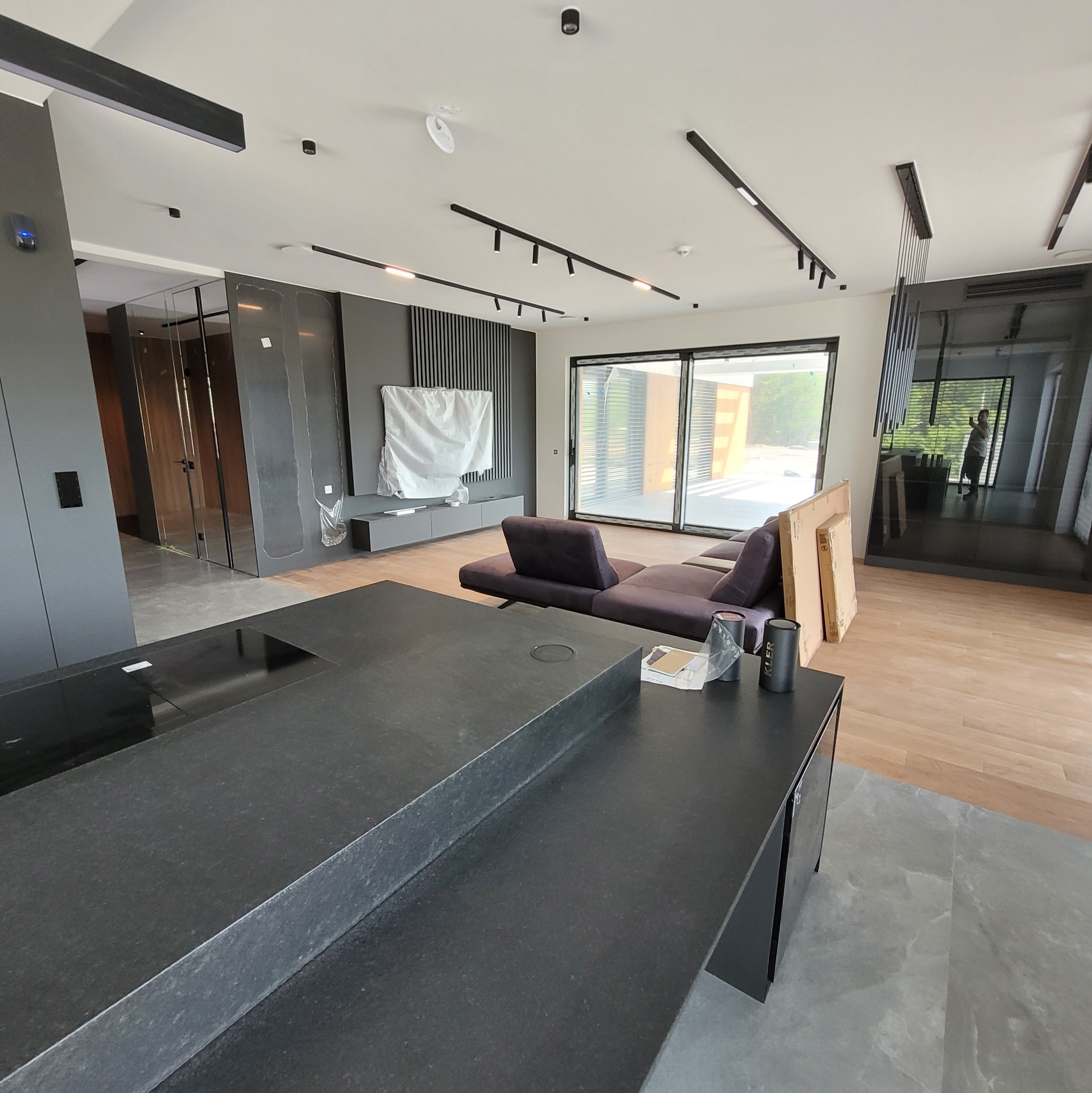 Nowoczesne wnętrze: ciemna zabudowa kuchenna z kamiennym blatem, salon z sofą i dużymi oknami, podłoga z jasnego drewna. Minimalistyczny design z akcentami czerni.