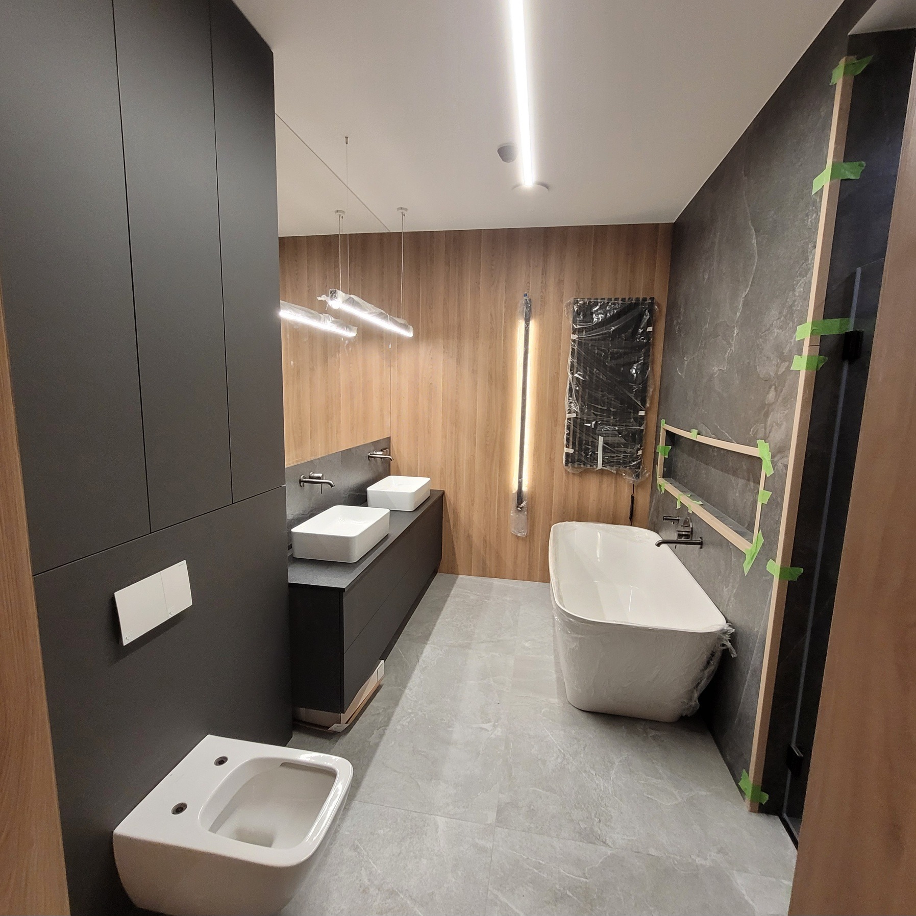 Nowoczesna łazienka w trakcie wykończenia: biała wanna i bidet, dwie umywalki na szafce, szare płytki podłogowe, drewniane i grafitowe panele ścienne, oświetlenie LED.