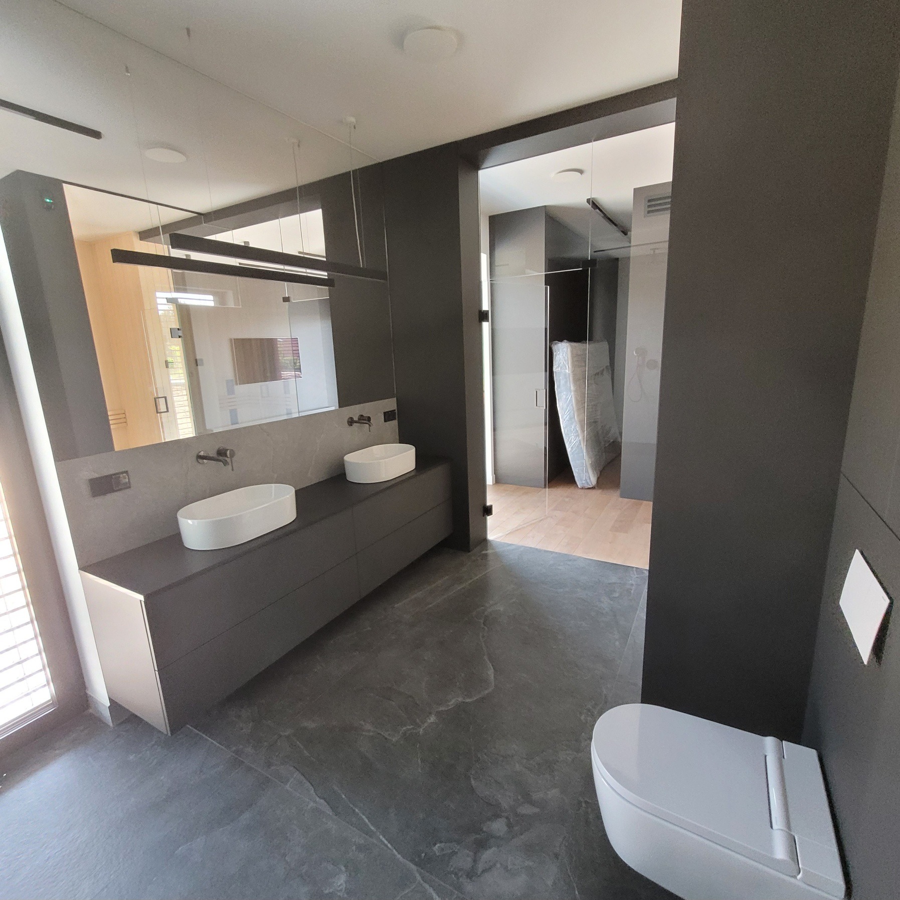 Nowoczesna łazienka z szarą armaturą, marmurową podłogą i minimalistycznym oświetleniem. Widoczny fragment toalety i wejście do innego pomieszczenia.