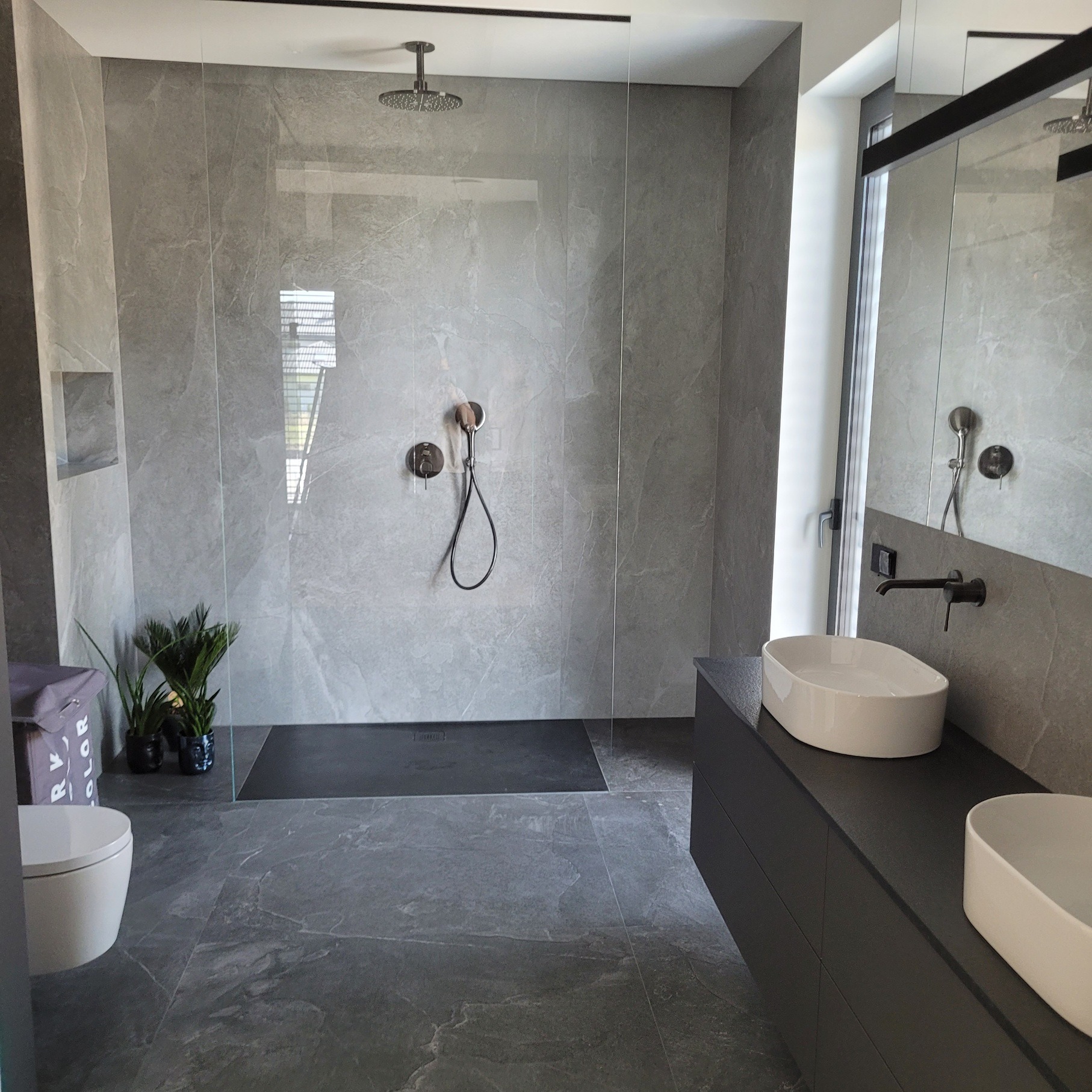 Nowoczesna łazienka z kabiną prysznicową typu walk-in, szarą armaturą i umywalkami nablatowymi. Minimalistyczny design, płytki imitujące kamień, czarny brodzik.