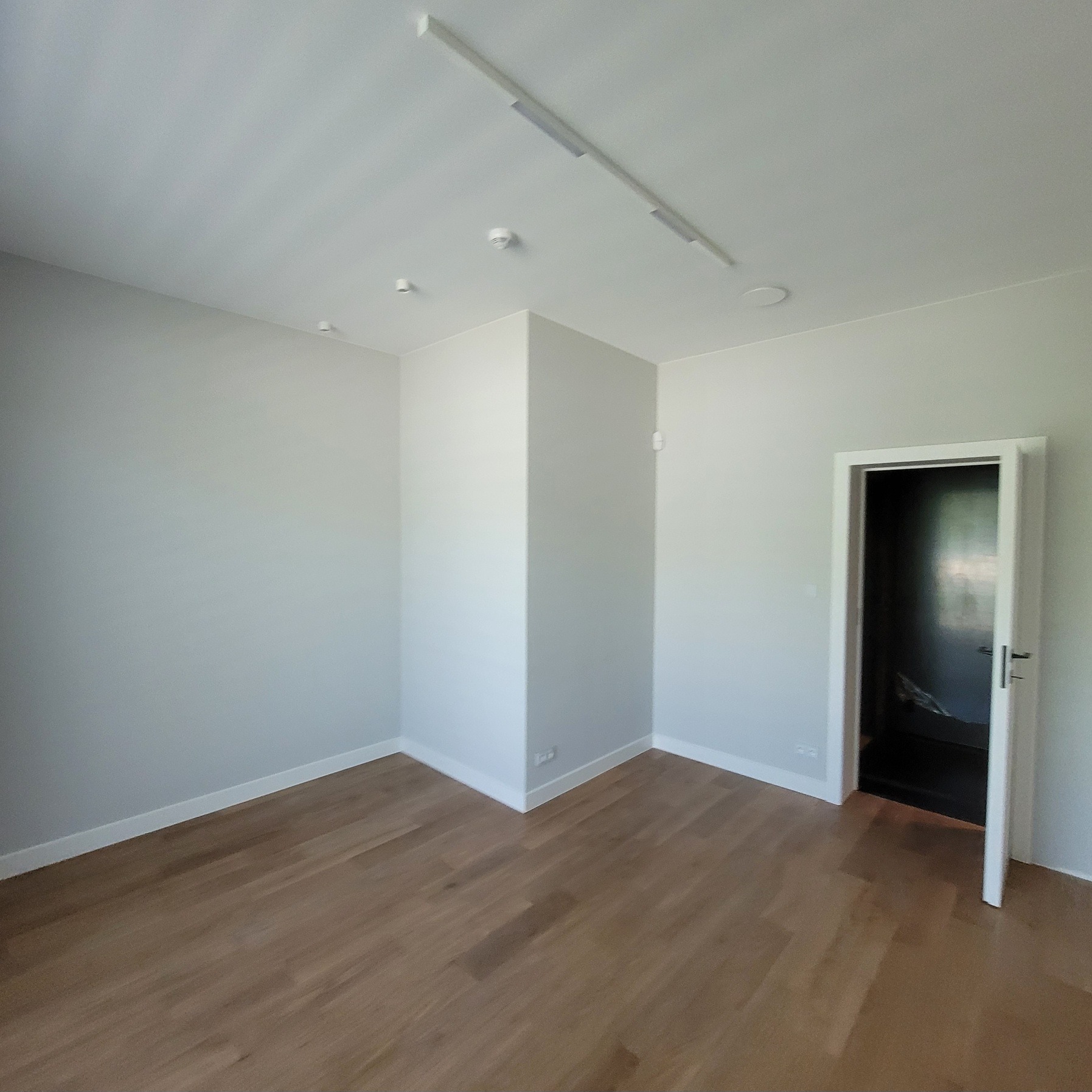 Jasne, puste wnętrze po remoncie: parkiet, białe ściany, listwy przypodłogowe i oświetlenie sufitowe. Otwarty pokój z drzwiami do innego pomieszczenia.