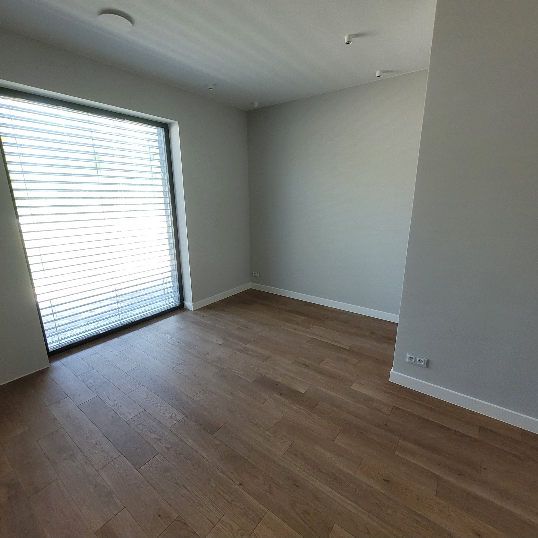 Jasne, wykończone wnętrze pokoju z drewnianą podłogą, szarymi ścianami i dużym oknem z żaluzjami. Widoczne listwy przypodłogowe i gniazdka elektryczne.